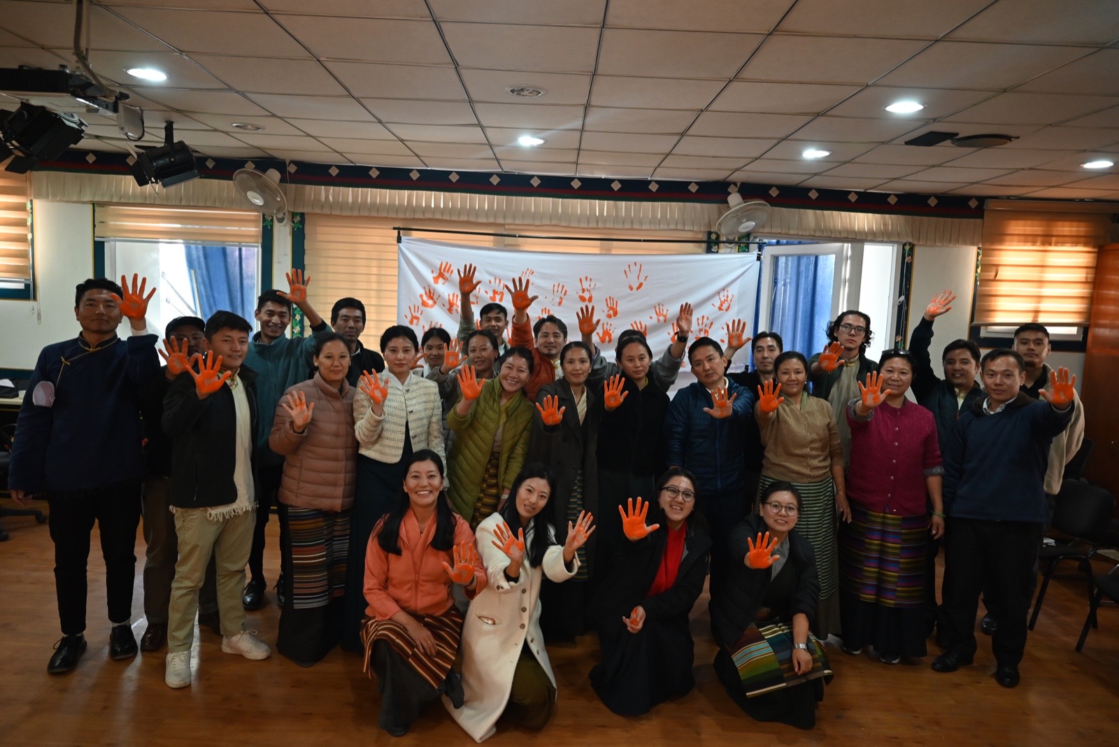 Group of people indoors posing for a workshop photo, all with orange-painted hands raised in the air, banner with orange handprints in the background.