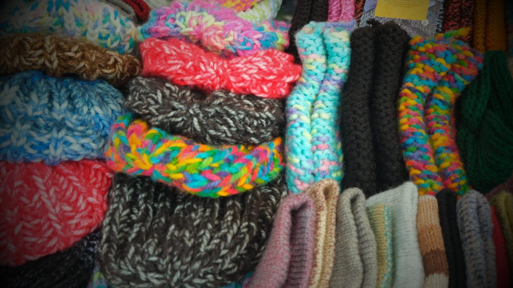 Pile of colorful knitted scarves and yarns stacked together in a market display, showing reds, blues, greens and rainbow hues.