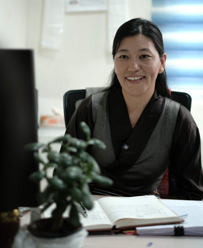 Smiling woman in a gray vest sits at a desk with an open notebook, plant, and computer monitor nearby.