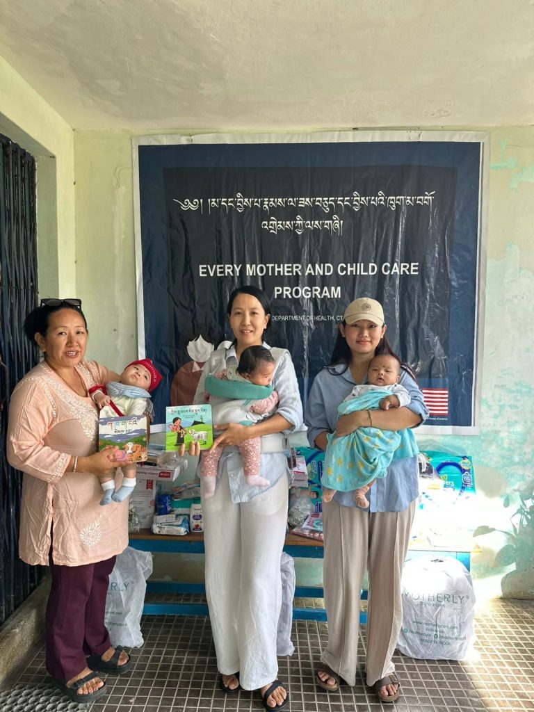 Three women holding babies in front of a banner that reads 'EVERY MOTHER AND CHILD CARE PROGRAM' with donated items on a table behind them.