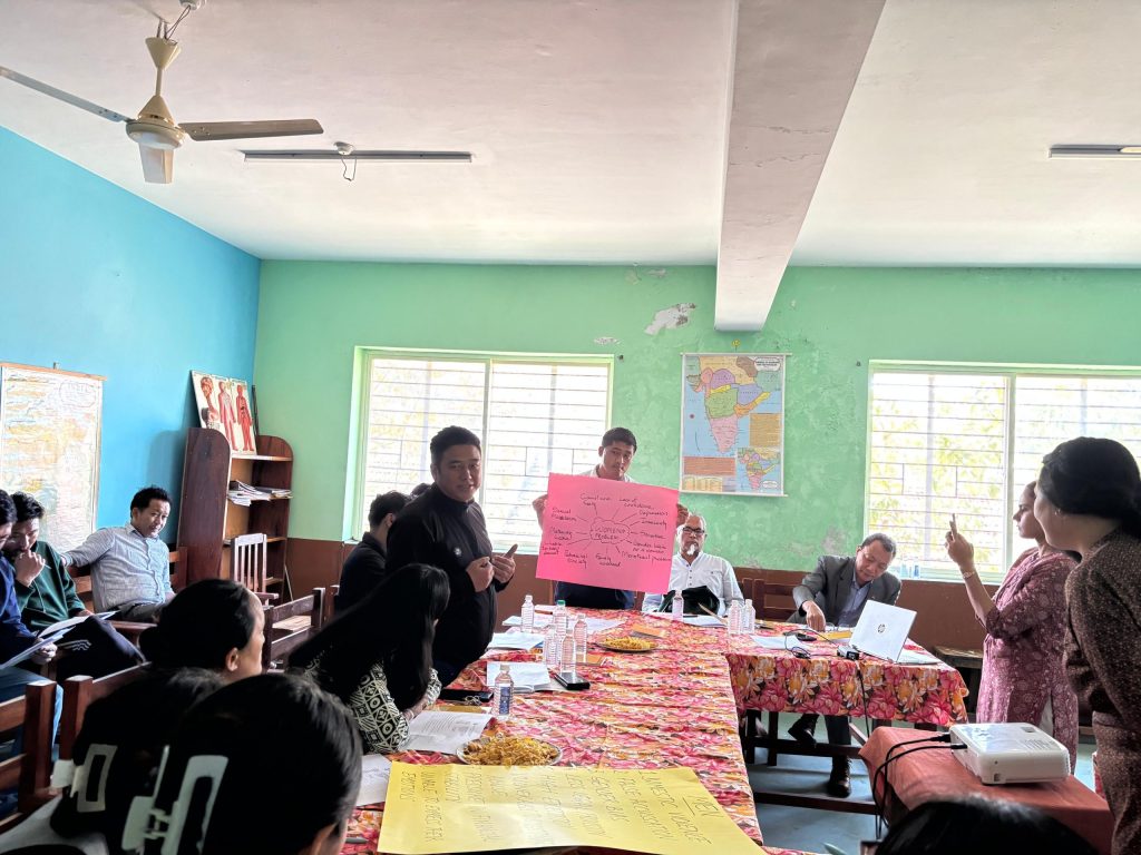 Adults in a bright classroom sit around tables, one person holds a pink poster while others discuss; maps and posters adorn the walls.