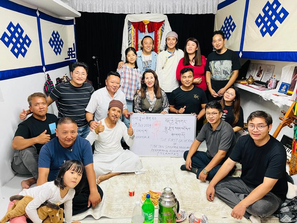 A diverse group of people sits and kneels on the floor in a room decorated with blue and white banners, posing for a group photo around a whiteboard with text.