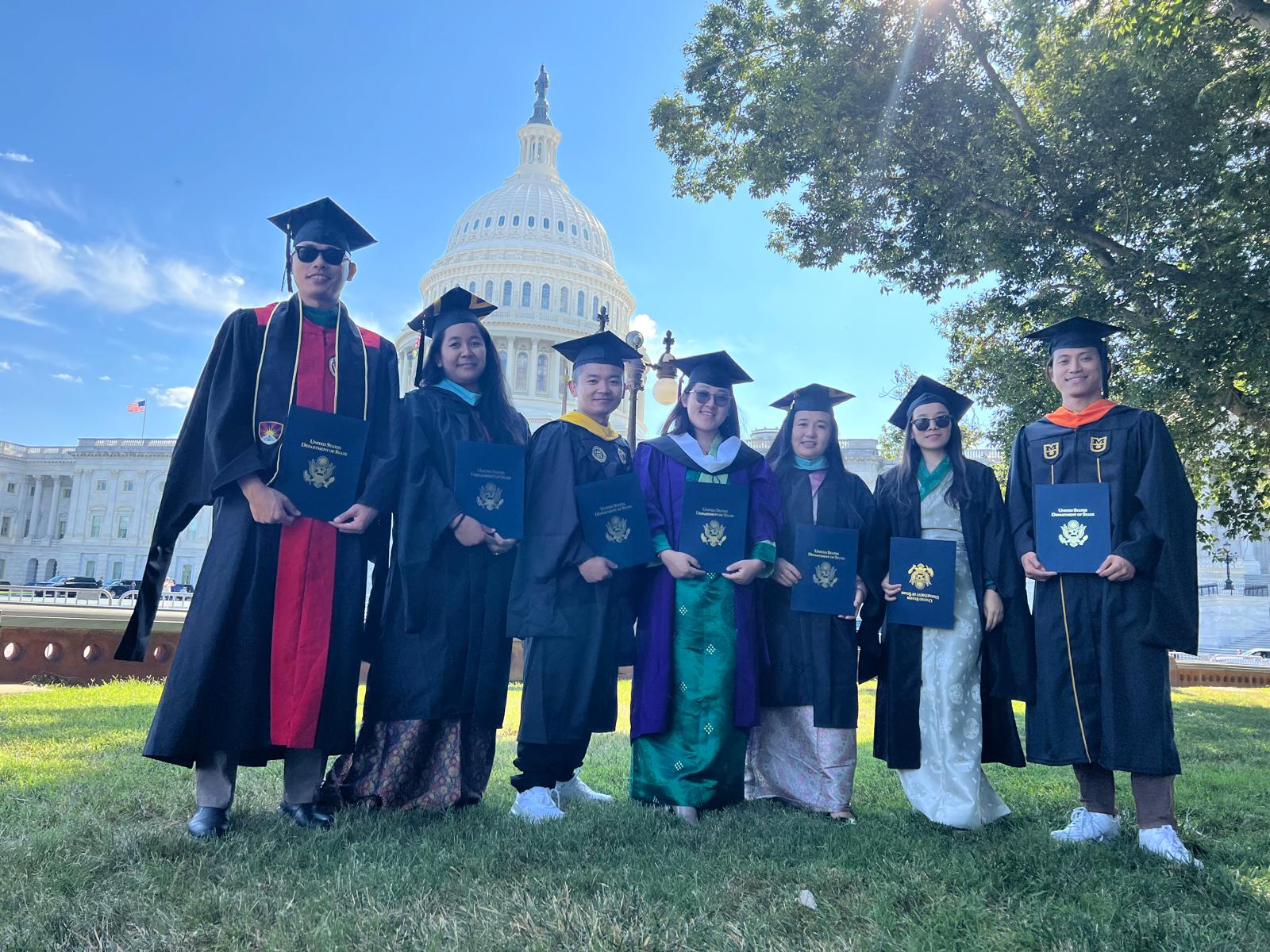 Group of graduates in caps and gowns posing on grass with the U.S. Capitol in the background on a sunny day.