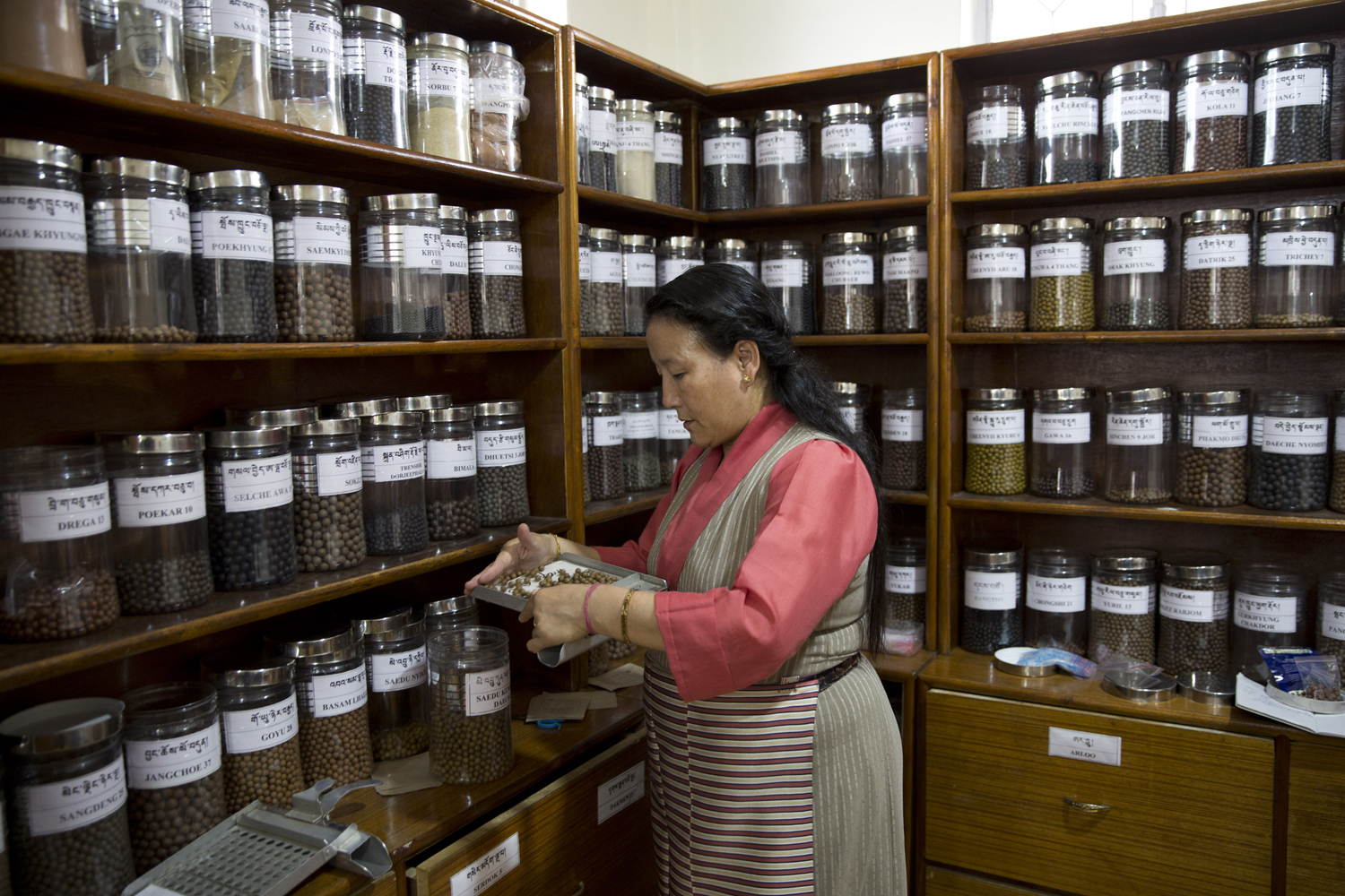 Woman in a shop with wooden shelves lined with labeled glass jars of dried seeds or beans, she’s measuring grains into a tray nearby