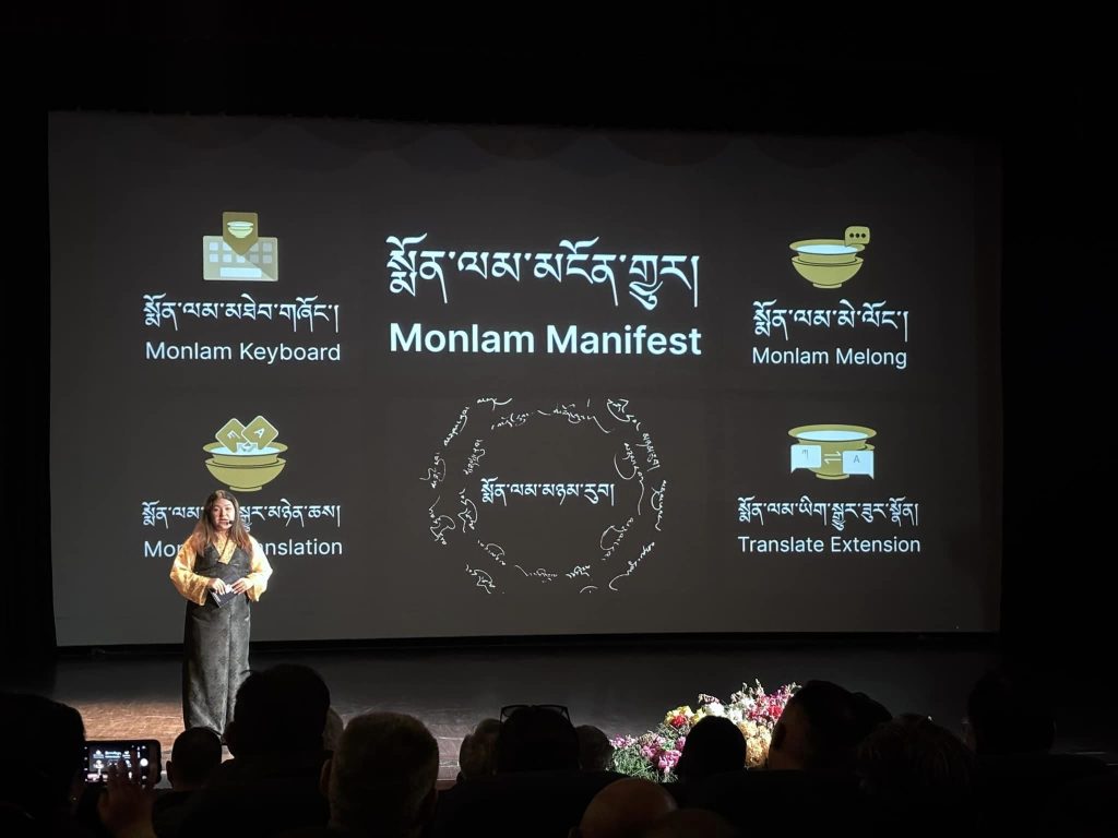 Presenter on a dark stage beside a large screen showing a slide titled 'Monlam Manifest' with icons labeled 'Monlam Keyboard' and 'Monlam Melong' and audience silhouettes in foreground.