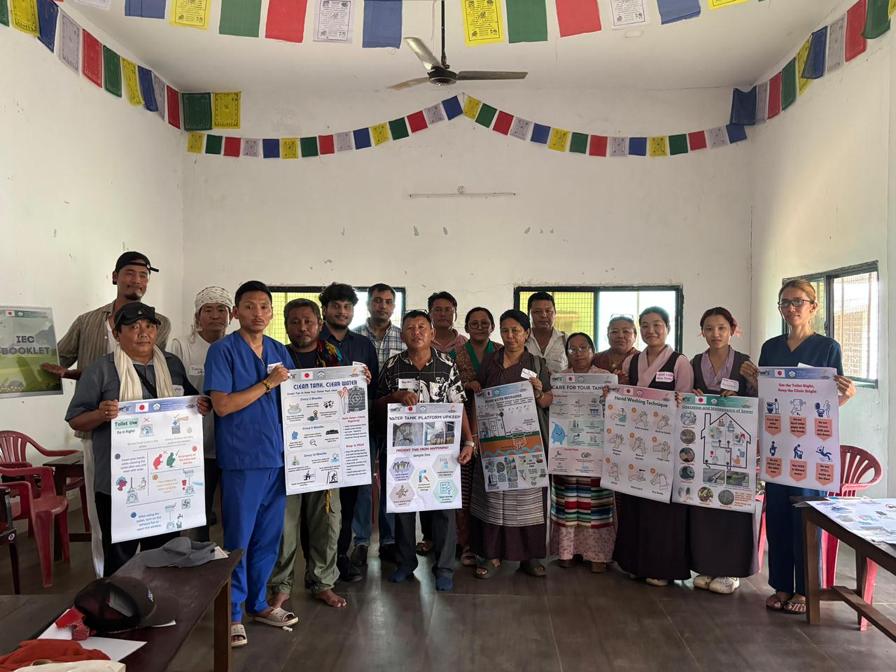 Group of about 18 adults standing in a bright room, each holding large informational posters about hygiene and safe water practices; colorful prayer flags hang from the ceiling.