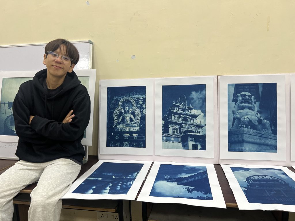 Young person in a black hoodie sits beside blue-toned art prints displayed on the wall and floor.