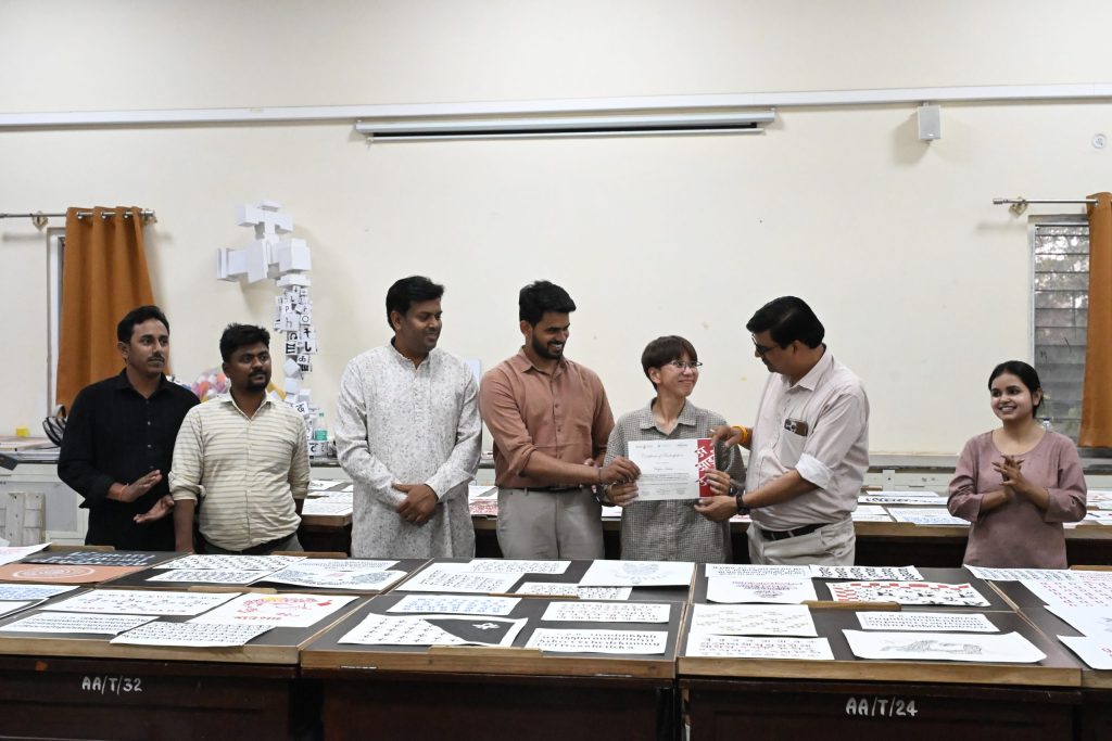 Group of people standing behind a table covered with printed sheets, one person receives a certificate amid smiles in a classroom setting.