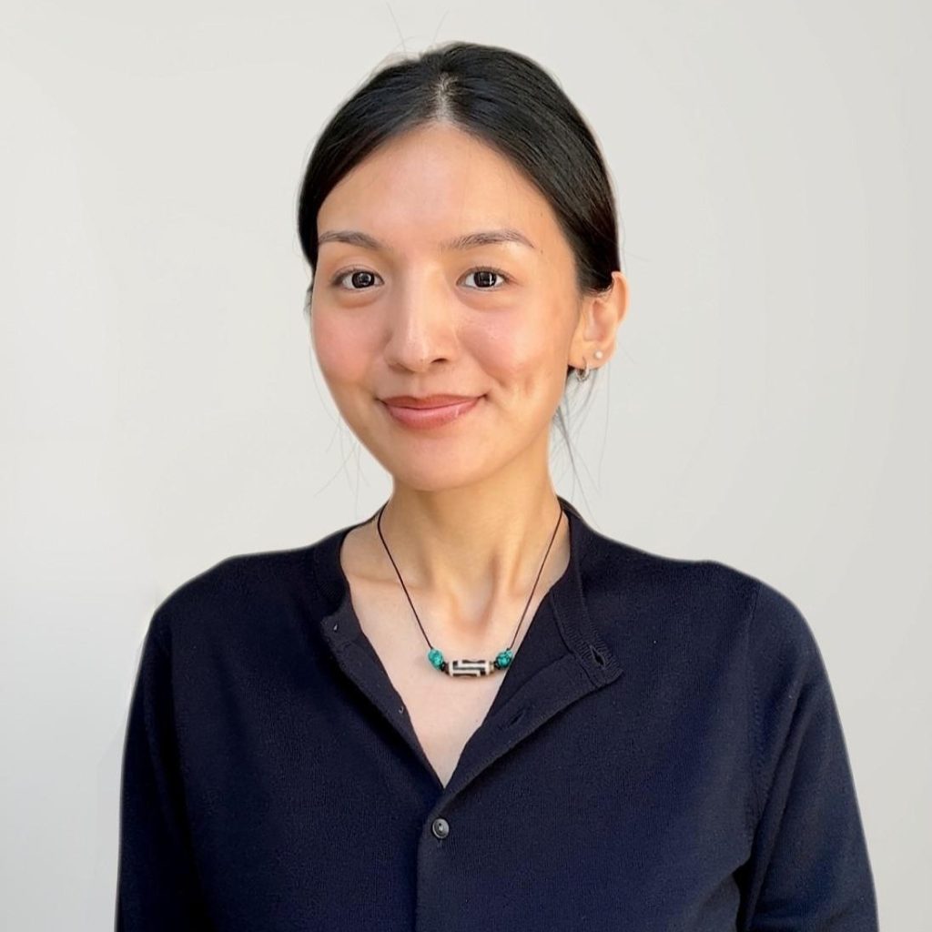Portrait of a smiling woman with dark hair, wearing a navy sweater and turquoise bead necklace, against a light gray background.