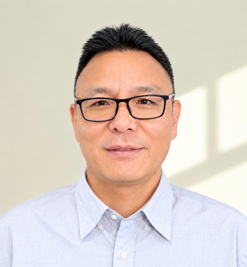 Portrait of a middle-aged man with short black hair and black-framed glasses wearing a light blue button-up shirt, neutral background