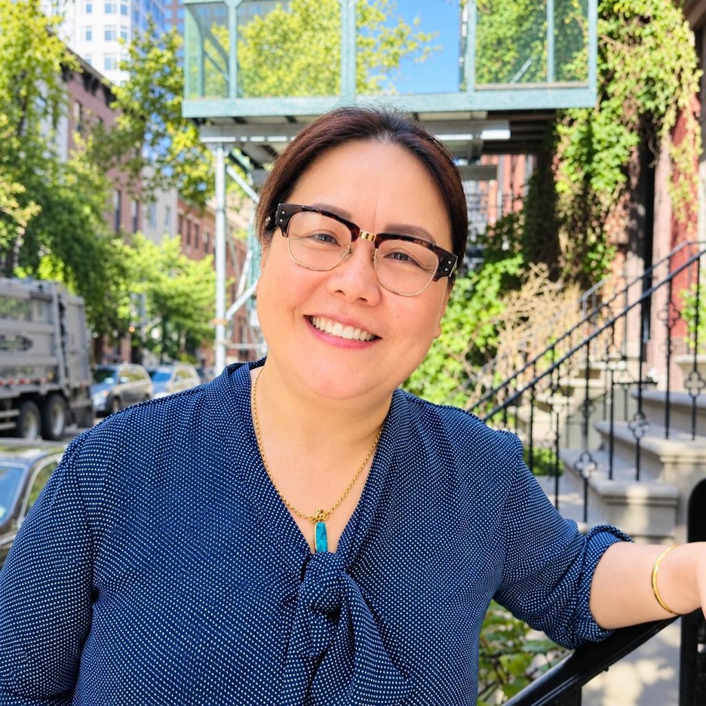 Smiling woman with glasses in a blue polka-dot blouse outdoors on a sunny city street.